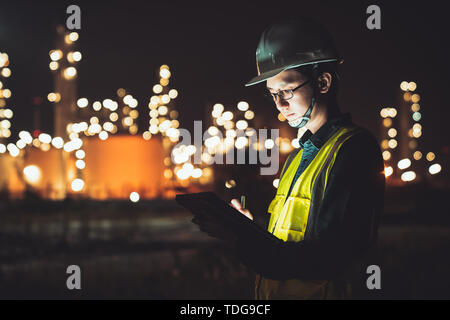 Asian man engineer using digital tablet working late night shift at petroleum oil refinery in industrial estate. Chemical engineering, fuel and power  Stock Photo