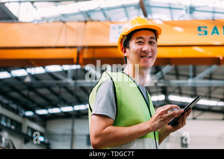 Young Asian male heavy industral worker holding a digital tablet inside manufacturing and factory site with crane behind Stock Photo