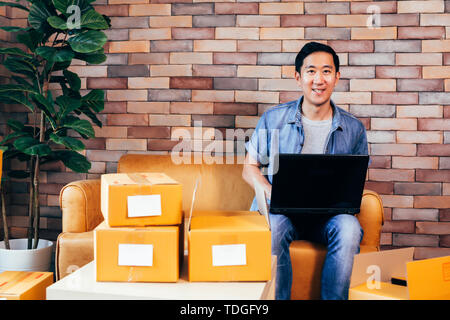 Young Asian male business entrepreneur using computer laptop while packing products into boxes to deliver - Online small business and ecommerce Stock Photo