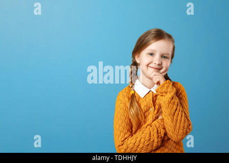 Portrait of a little girl schoolgirl in a yellow sweater on a blue background. Pensive child holding hand on chin and looking at camera. Copy space Stock Photo