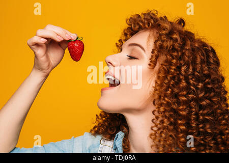 Smiling curly red-haired woman wearing chinese hat holding photo camera and making photo on yellow background. Tourism and travel Stock Photo