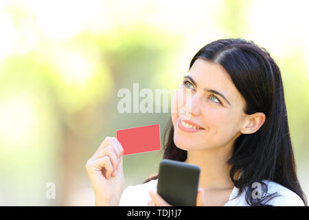 Happy online shopper holding smart phone showing blank credit card and looking at side in a park Stock Photo