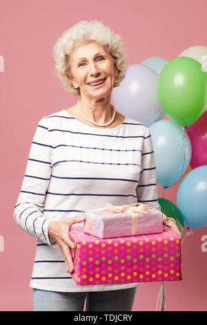 Cheerful optimistic curly-haired mature lady with necklace wearing stripped shirt standing against heap of balloons and holding gifts while enjoying b Stock Photo