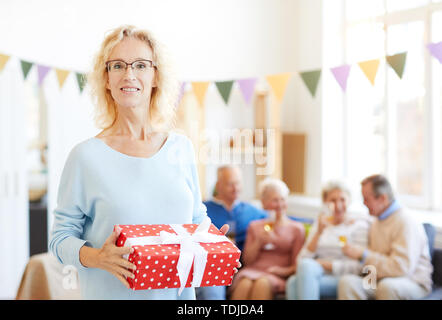 Portrait of positive attractive mature lady with curly hair holding big gift box wrapped in ribbon while her friends chatting and drinking alcohol in  Stock Photo