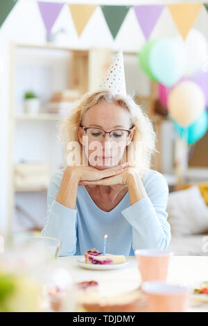 Depressed beautiful mature lady in party hat sitting at table and leaning on hands while thinking about birthday wish Stock Photo