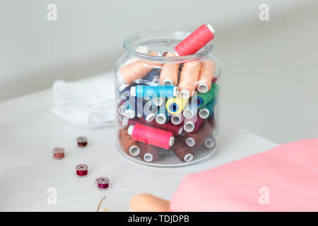 Close up multicolored threads and bobbins lie on a table in a transparent glass jar on the seamstress's desktop. The concept of unique designer Stock Photo