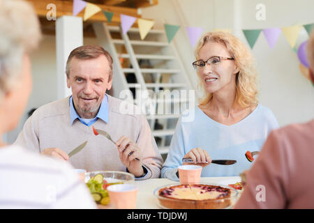 Positive mature friends in casual sweaters sitting at table and eating food while discussing news at dinner party Stock Photo
