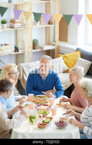 Content satisfied mature man in blue sweater sitting at table and listening to news of friends during dinner party, occasion to gather friends Stock Photo