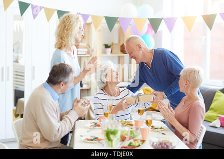 Group of positive excited people celebrating birthday of retired pretty woman at home, handsome man giving gift to birthday woman Stock Photo