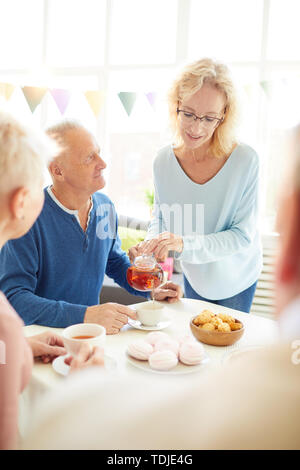 Positive attractive woman in glasses standing at dining table and pouring tea for guest while they talking together at party Stock Photo