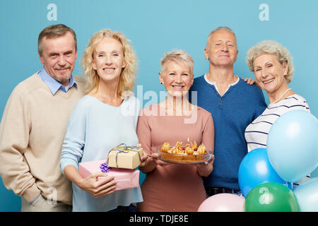 Cheerful mature friends gathering for birthday party of lady holding sweet cake, they posing together against blue background Stock Photo