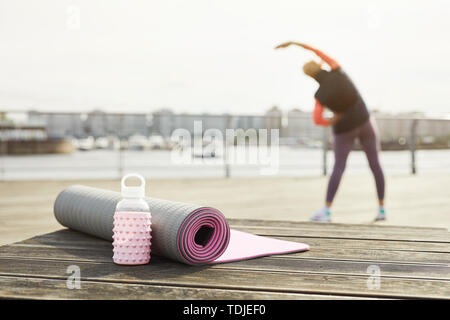 Back view of unrecognizable woman stretching on wooden pier outdoors with focus on yoga mat in foreground, copy space Stock Photo