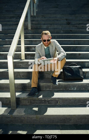 Serious concentrated hipster businessman with red beard sitting on stairs outdoors and working with tablet Stock Photo