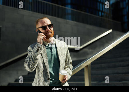 Content confident handsome white-collar man with read beard standing on stairs and drinking coffee while talking on phone Stock Photo