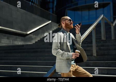 Content confident fashionable male entrepreneur with red beard leaning on railing outdoors and recording voice message on smartphone Stock Photo