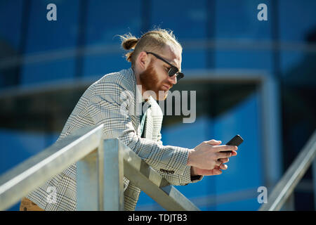 Serious busy hipster man in sunglasses leaning on railing and using smartphone while texting sms outdoors Stock Photo
