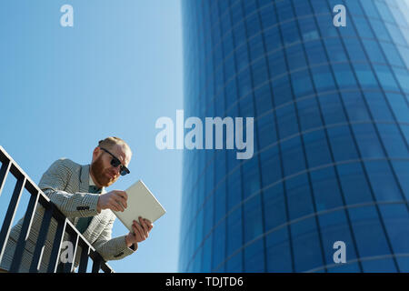 Below view of serious city guy in sunglasses leaning on railing in financial district and working with digital tablet Stock Photo