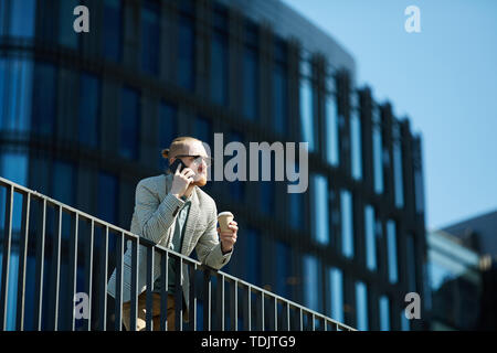 Serious pensive freelance manager with red beard leaning on railing and contemplating financial district while talking to customer on phone Stock Photo