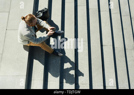 Above view of pensive stylish businessman with ponytail sitting on stairs and surfing net on smartphone Stock Photo