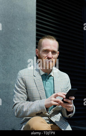 Smiling red-bearded manager in stylish jacket sitting outdoors and texting sms on smartphone while looking at camera Stock Photo