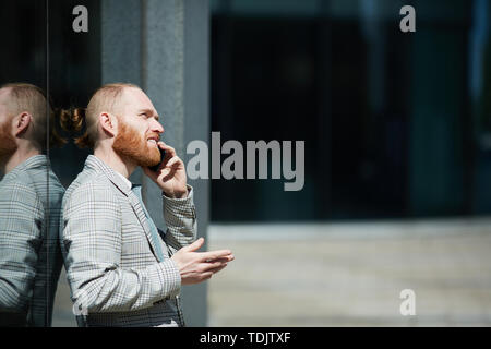 Thoughtful handsome hipster entrepreneur with beard leaning on wall outdoors and gesturing hand while discussing deal on phone Stock Photo