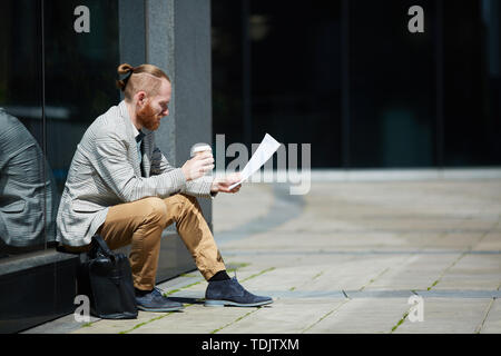 Thoughtful handsome young freelance analyst sitting outdoors and examining data report while drinking coffee Stock Photo