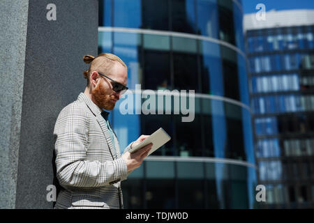 Serious thoughtful hipster young manager in sunglasses standing in business district and browsing mobile internet on tablet Stock Photo