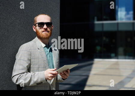 Portrait of content confident hipster businessman in sunglasses standing by gay wall and using tablet for work Stock Photo