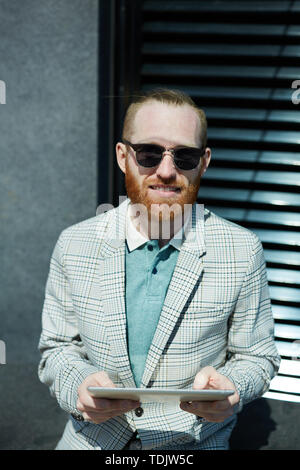 Portrait of cheerful hipster handsome young businessman in sunglasses using digital tablet on move Stock Photo