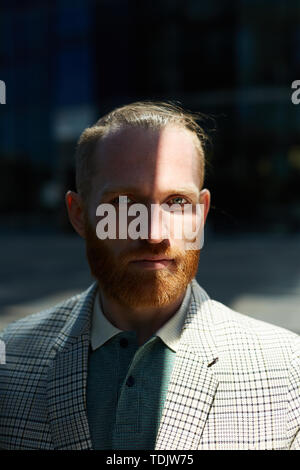 Portrait of serious confident businessman with red mustache and beard wearing fashionable checkered jacket standing in shade outdoors Stock Photo