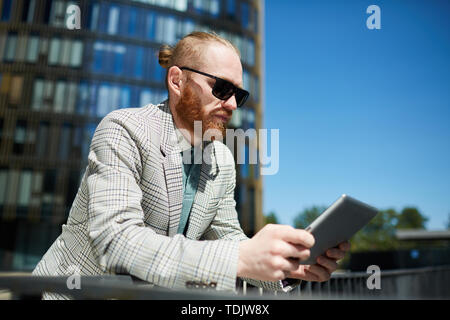 Serious thoughtful young bearded manager in sunglasses leaning on railing in city and viewing information on tablet Stock Photo