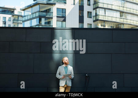 Content determined businessman with red beard standing by wall outdoors and using tablet while finding inspiration for project Stock Photo