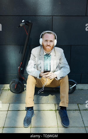 Smiling handsome young office employee in wired headphones sitting on deck of kick scooter and using mobile app of gadget Stock Photo