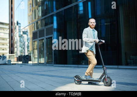 Portrait of positive active man with beard riding push scooter while commuting home after work day Stock Photo