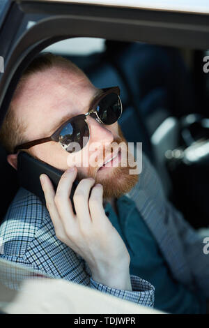 Smiling pensive hipster bearded businessman in sunglasses searching for location while talking on phone inside of car Stock Photo