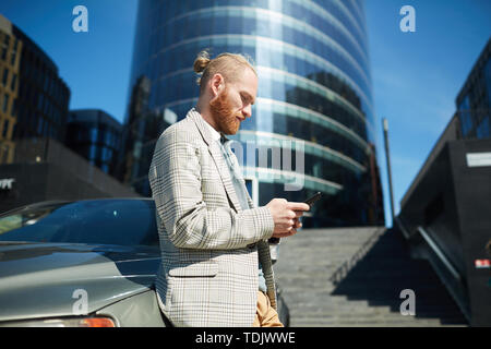 Serious thoughtful hipster young businessman with ponytail leaning on car outdoors and texting message on phone Stock Photo