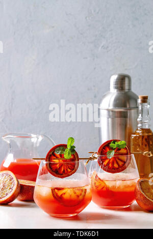 Blood orange iced cocktails in glasses, decorated by slice of oranges and fresh mint on skewers, served with shaker, jug of juice, bottle of rum on wh Stock Photo