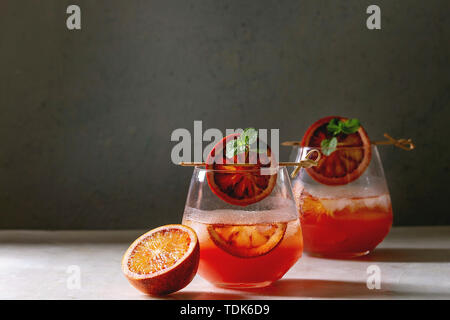 Blood orange iced cocktails in glasses, decorated by slice of oranges and fresh mint on skewers, served on white marble table with grey wall at backgr Stock Photo