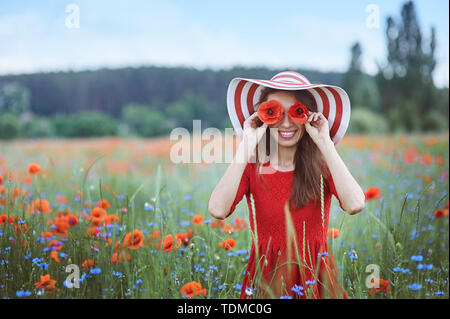 Beautiful young woman holding red poppies in front of her eyes and standing in flower meadow Stock Photo