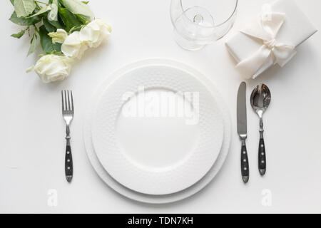 White table setting with bouquet rose, gift, dishware, silverware on holiday white table. Top view. Romantic and wedding anniversary. Stock Photo