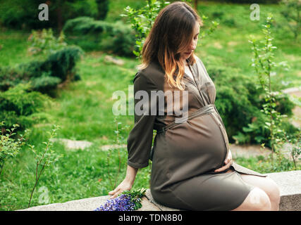 Pregnant woman sitting in the park looking at her belly. Green grass background. Stock Photo