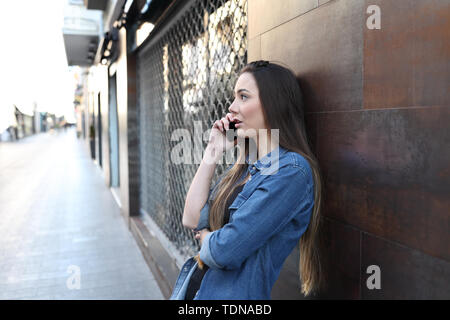 Serious woman talks on smart phone leaning in a wall in the street Stock Photo