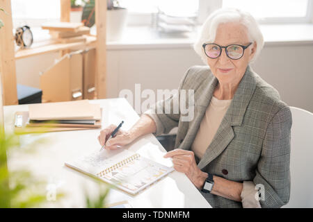 Elderly businesswoman with white hair writing down plan of working day in notebook by desk in front of camera Stock Photo