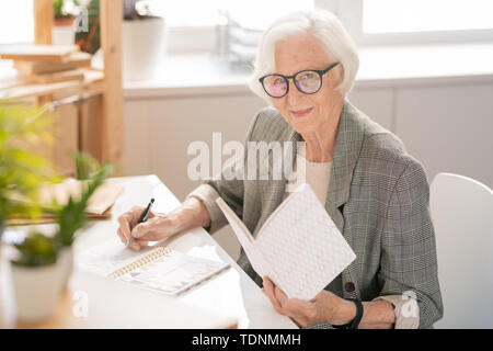 Happy senior white-haired businesswoman with open book making notes in notebook while reading by desk Stock Photo