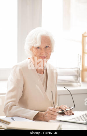 Aged successful female business leader in white elegant suit sitting by workplace in front of laptop during work Stock Photo