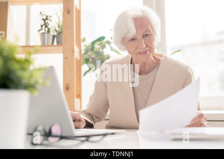 Aged female economist or banker looking through financial papers while sitting by desk in front of laptop in office Stock Photo