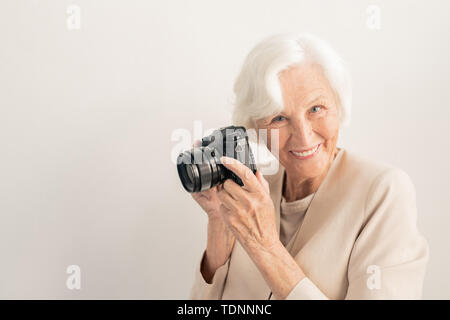 Successful senior female photographer with camera looking at you with toothy smile over white background in isolation Stock Photo