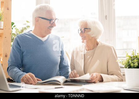 Happy senior couple in smart casualwear looking at each other while sitting by desk and looking through magazine Stock Photo