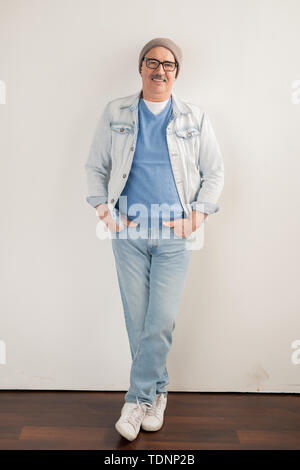 Happy mature man in jeans, pullover, jacket, beanie and sneakers leaning over white wall in studio while looking at you Stock Photo