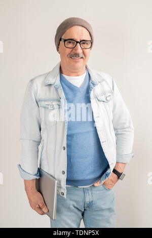 Casual mature man in blue jeans and pullover, white jacket, eyeglasses and beanie standing in front of camera over white background Stock Photo